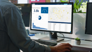 A city worker at a desktop computer displaying the Urban Hawk infrastructure management dashboard with pavement detection analytics. The screen features a map highlighting roadway conditions, charts summarizing detections and lane miles covered, and a table of inspection data, illustrating how digital tools help monitor and prioritize road maintenance.